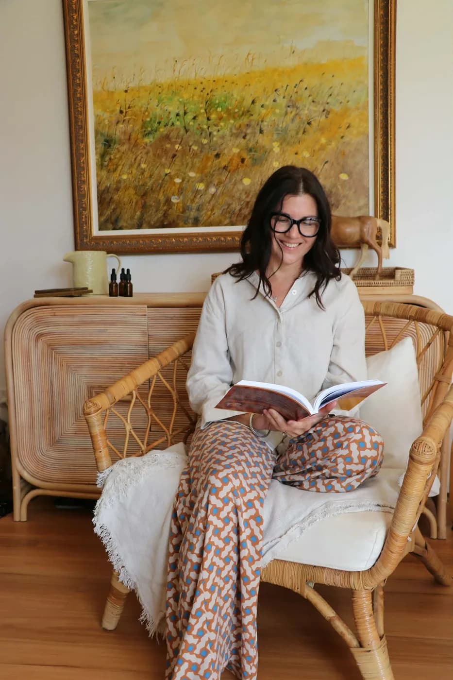 Celeste Langridge reading in a cane chair, amber remedy bottles on the sideboard behind her
